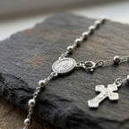 Close-up of silver Miraculous Medal, spring ring clasp, polished bead chain and decorative crucifix resting on dark stone.