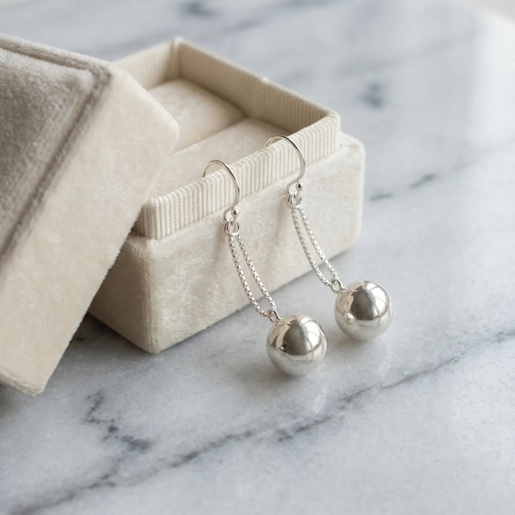 Silver sphere drop earrings draped over open Luna Dura cream velvet gift box on white marble surface Dubai Luna Dura Studio
