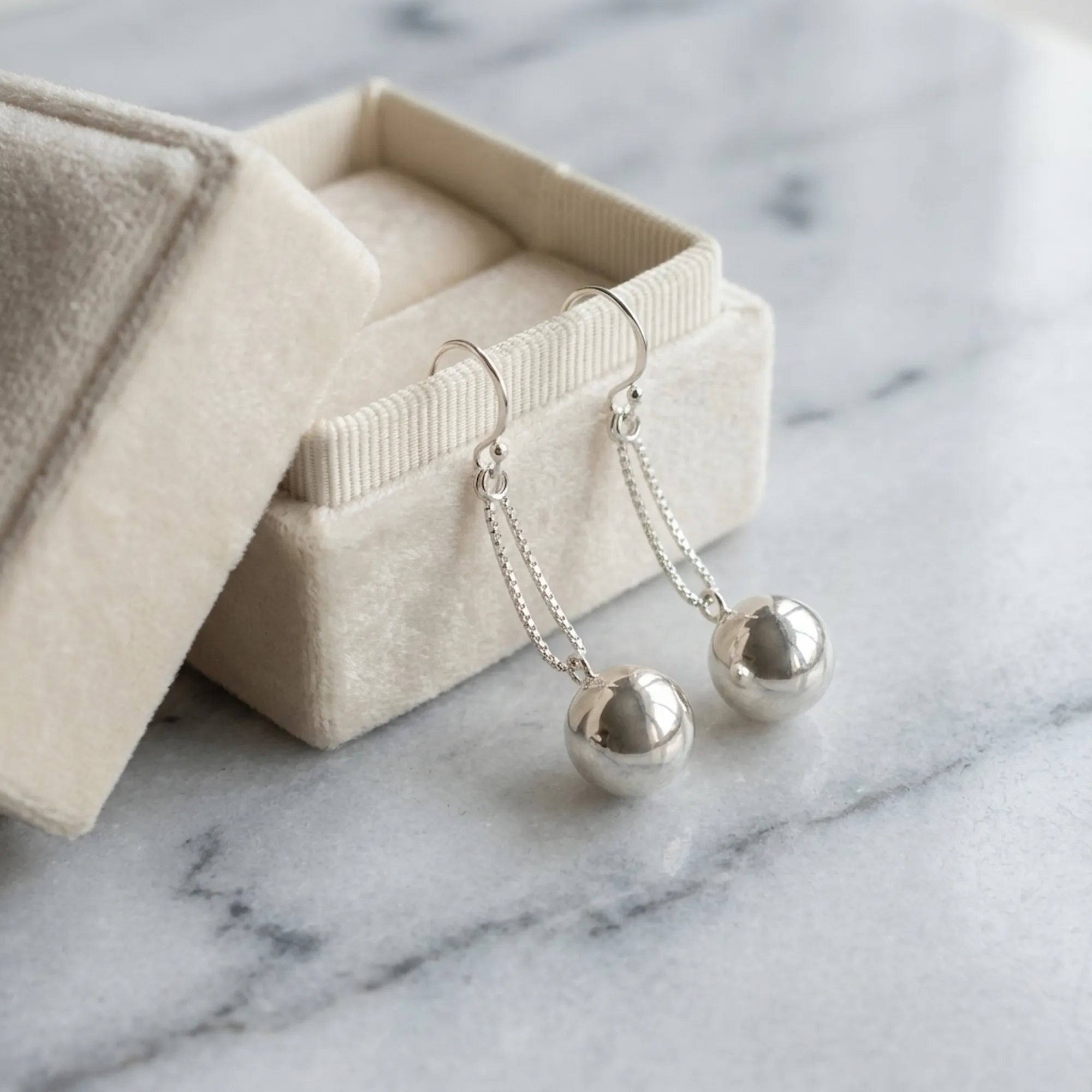 Silver sphere drop earrings draped over open Luna Dura cream velvet gift box on white marble surface Dubai Luna Dura Studio