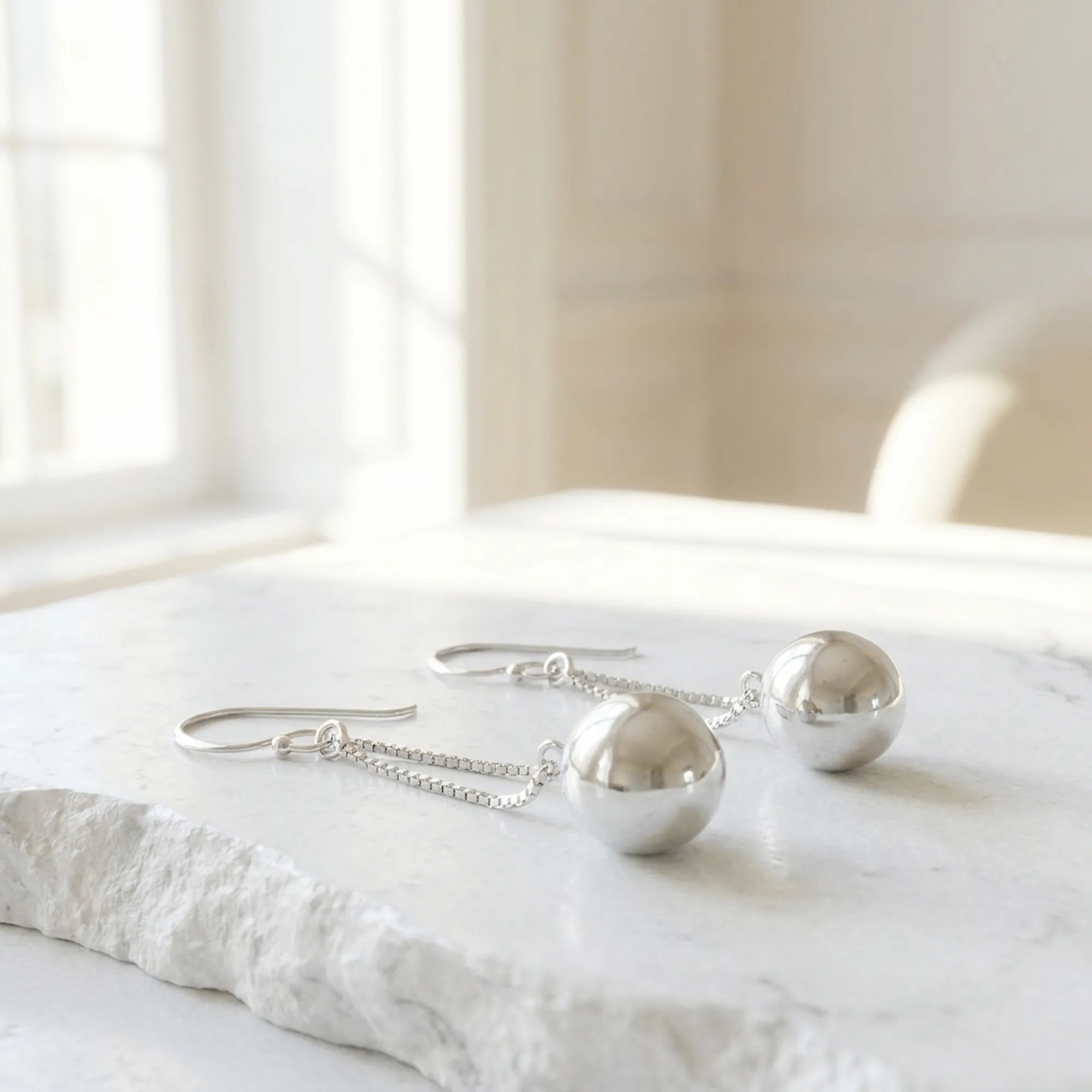 Pair of silver sphere drop earrings laid on white stone surface in bright minimal interior setting UAE.
