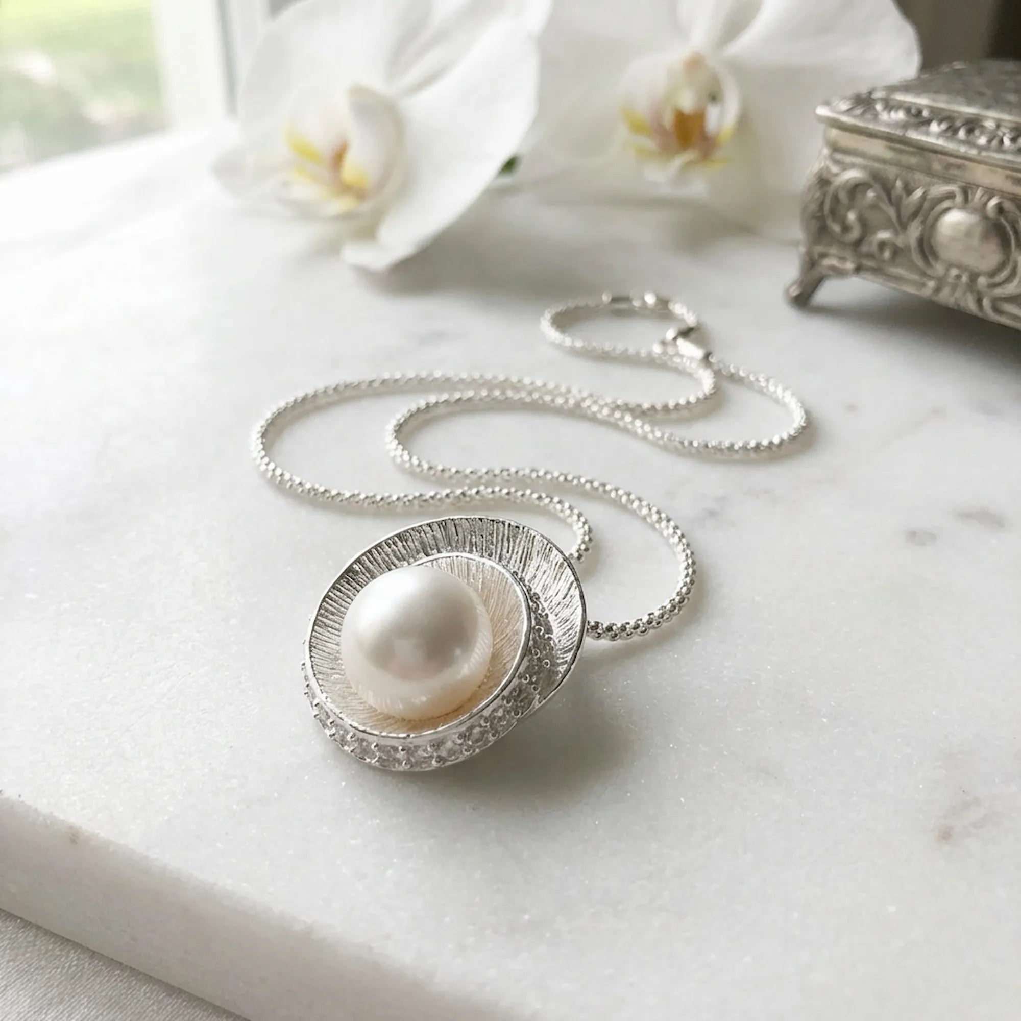 Spiral pearl halo necklace laid flat on marble with orchids and ornate silver jewellery box