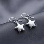 Pair of polished silver star drop earrings resting on dark grey fabric, showing three-dimensional puffed silhouette