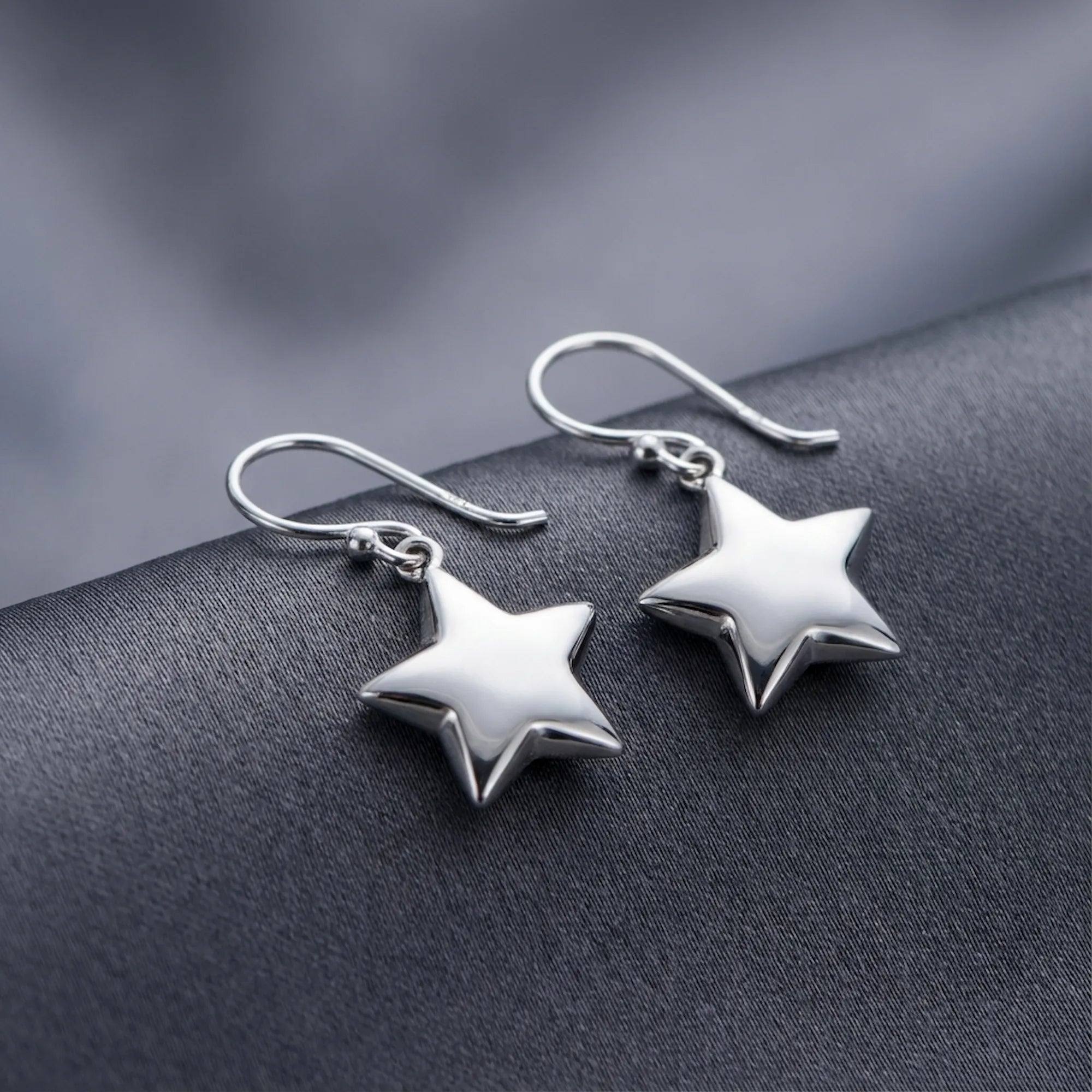 Pair of polished silver star drop earrings resting on dark grey fabric, showing three-dimensional puffed silhouette