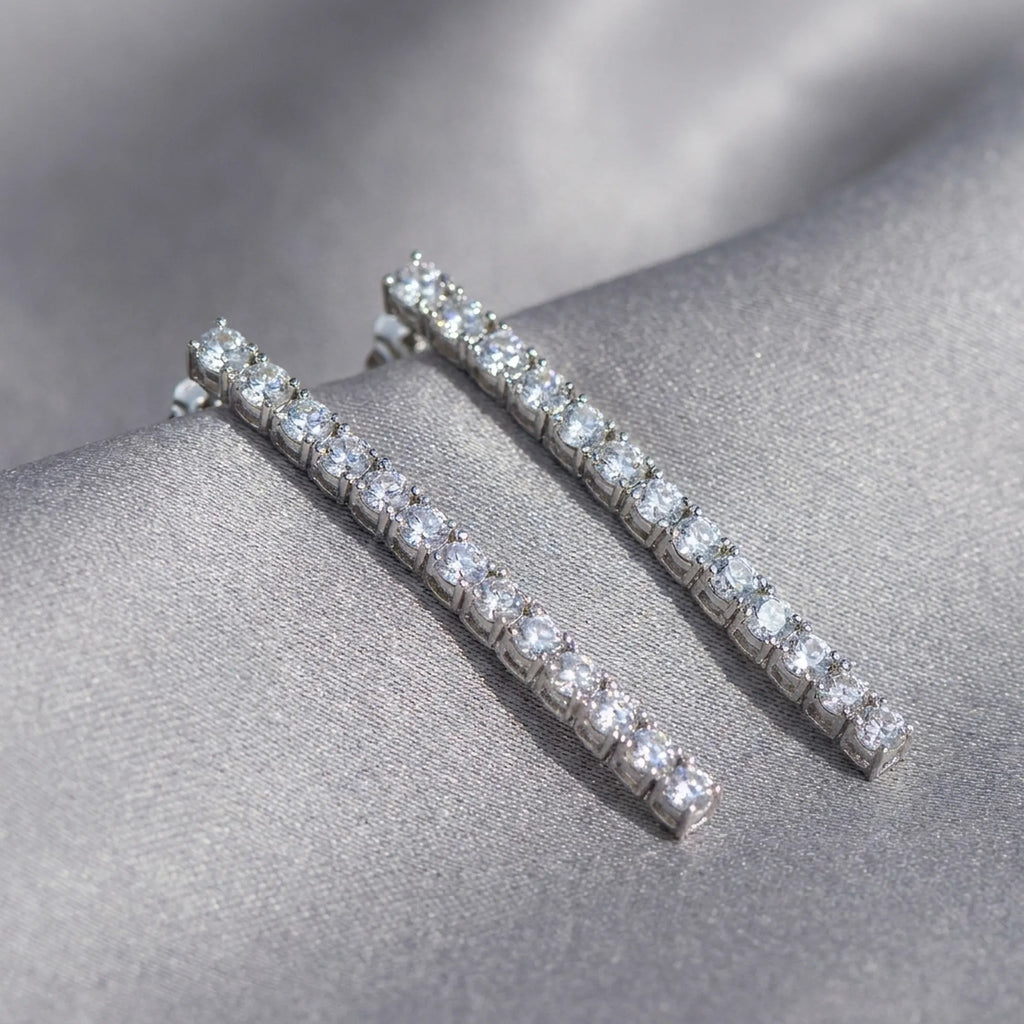 Tennis drop earrings flat lay on grey satin — individual prong-set CZ stones visible across full 40mm line rhodium silver