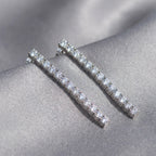 Tennis drop earrings flat lay on grey satin — individual prong-set CZ stones visible across full 40mm line rhodium silver