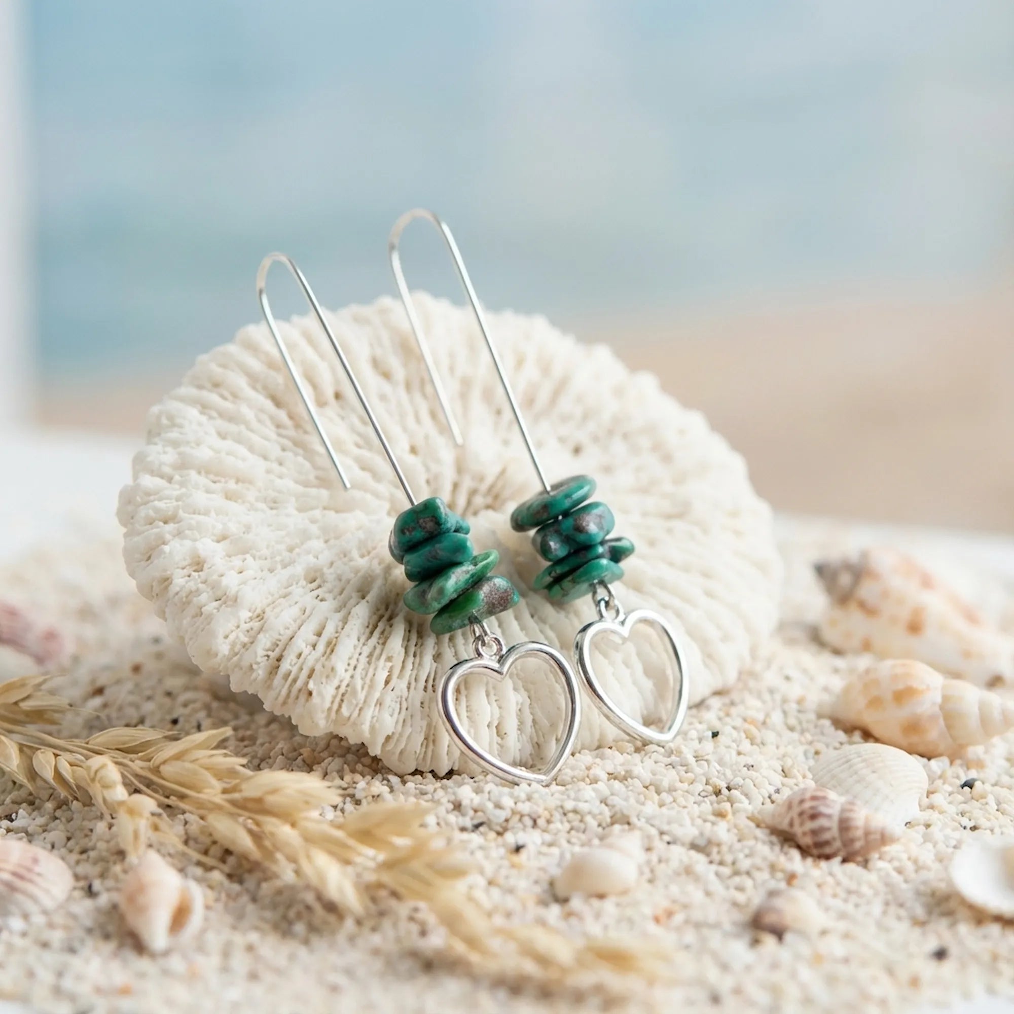 Raw turquoise and silver heart drop earrings on white coral in coastal sand setting — seashells and dried grass