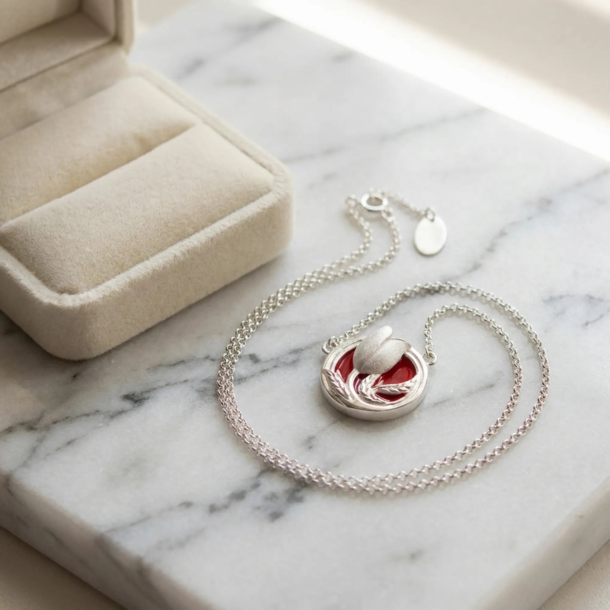Red enamel tulip medallion necklace laid on marble beside cream velvet jewelry box