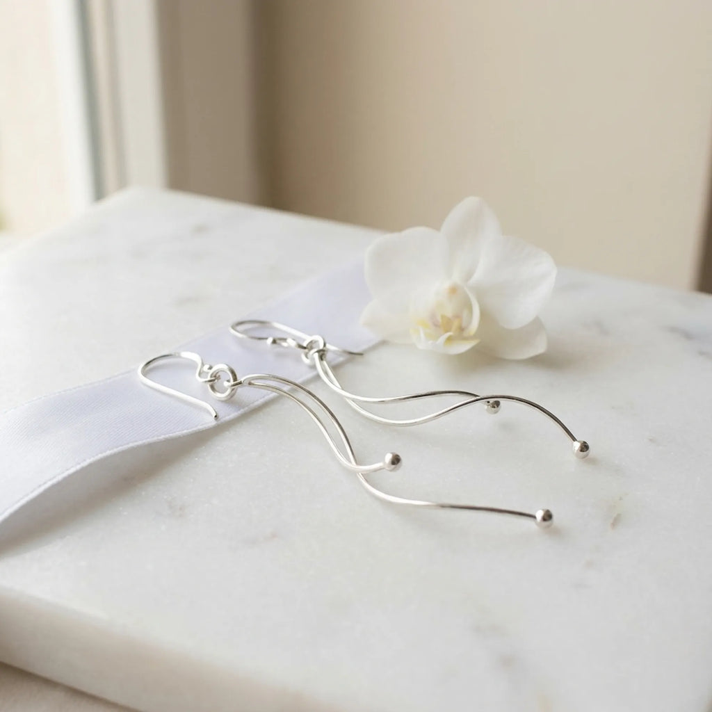 Silver wave drop earrings on marble surface with white orchid and satin ribbon, bright polish finish