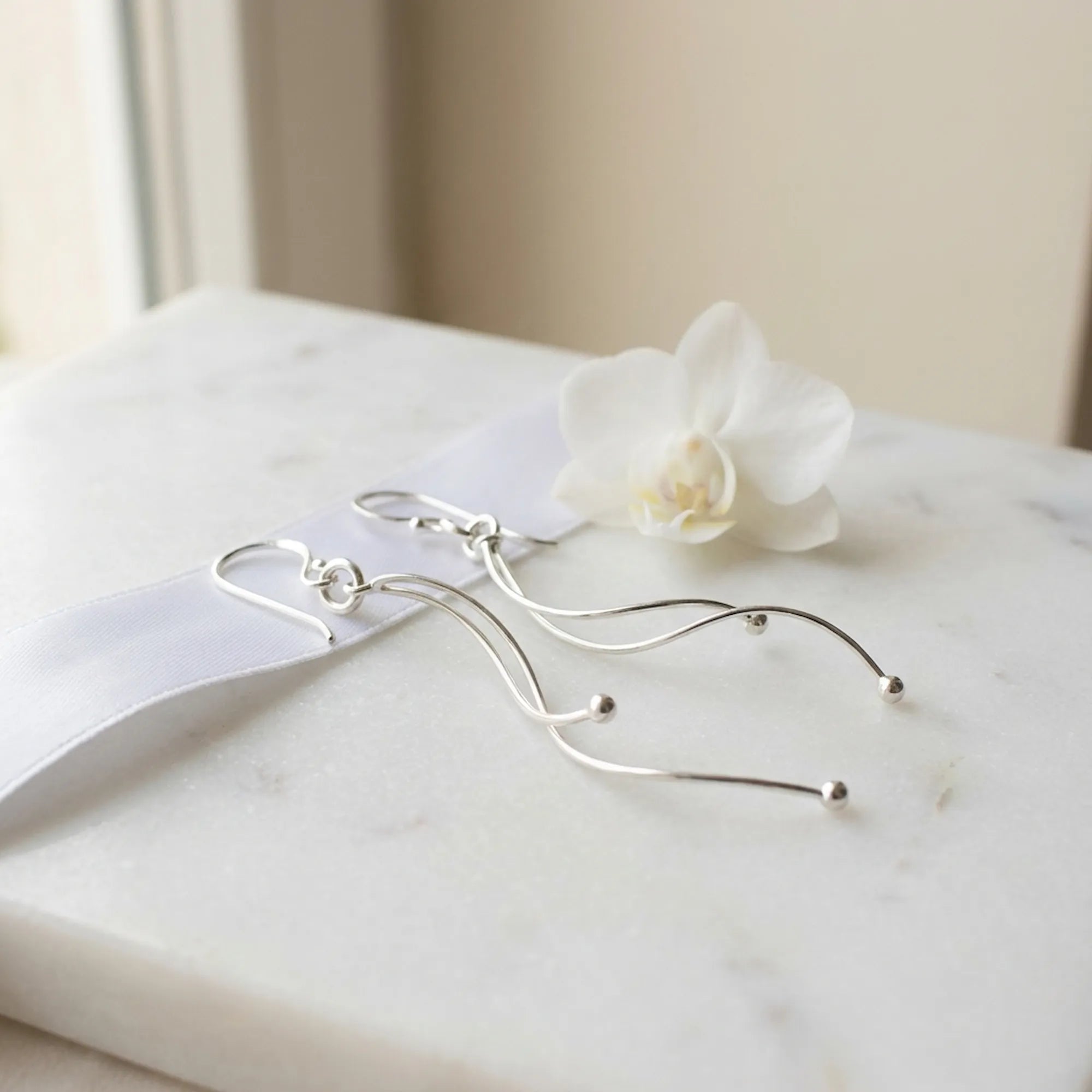 Silver wave drop earrings on marble surface with white orchid and satin ribbon, bright polish finish