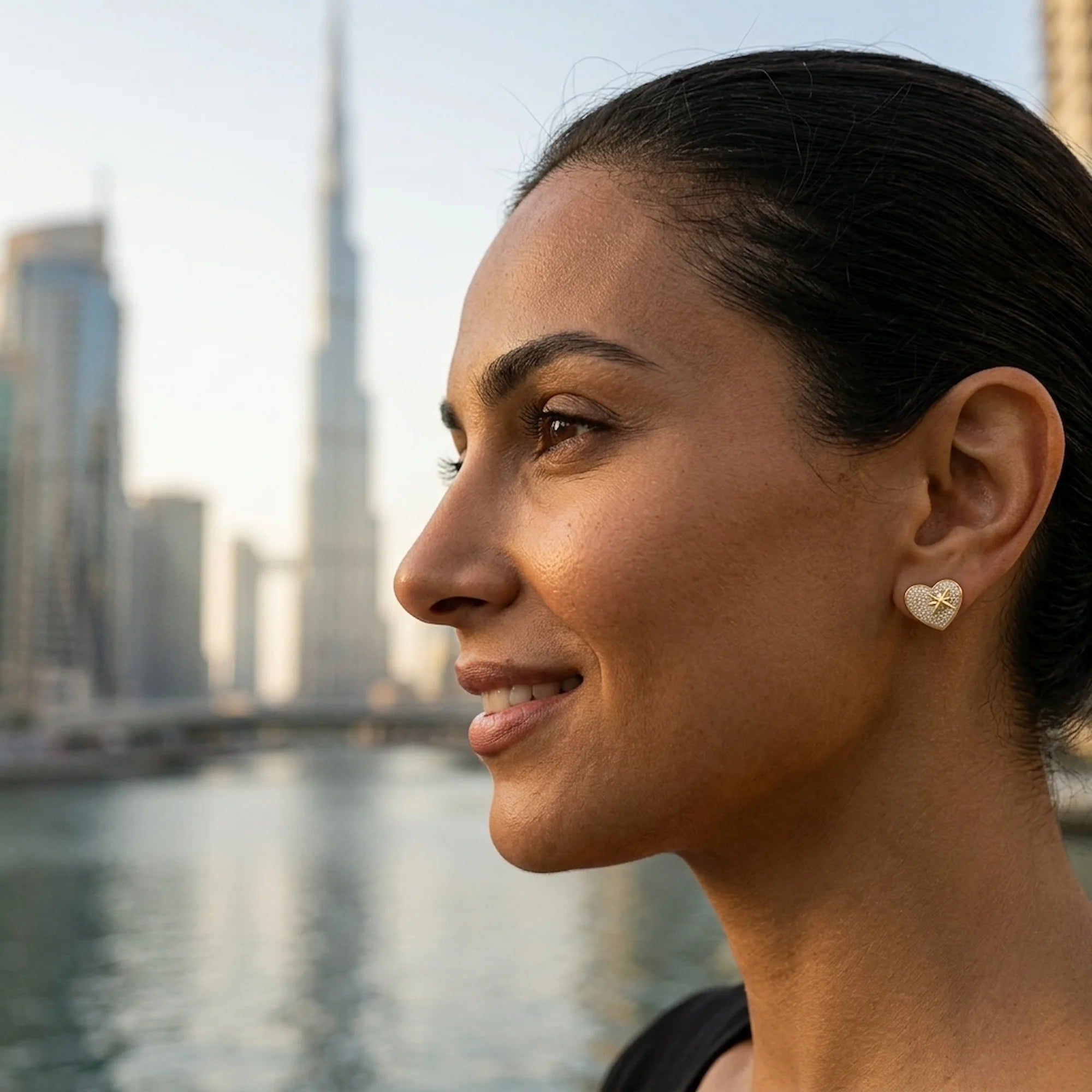 Woman wearing yellow gold 925 silver pavé heart earrings with cubic zirconia in Dubai near Burj Khalifa.