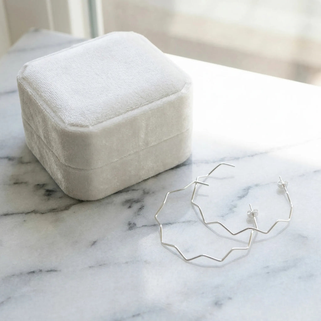 Silver zigzag geometric hoop earrings beside a cream velvet gift box on marble. Luna Dura.