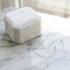 Silver zigzag geometric hoop earrings beside a cream velvet gift box on marble. Luna Dura.